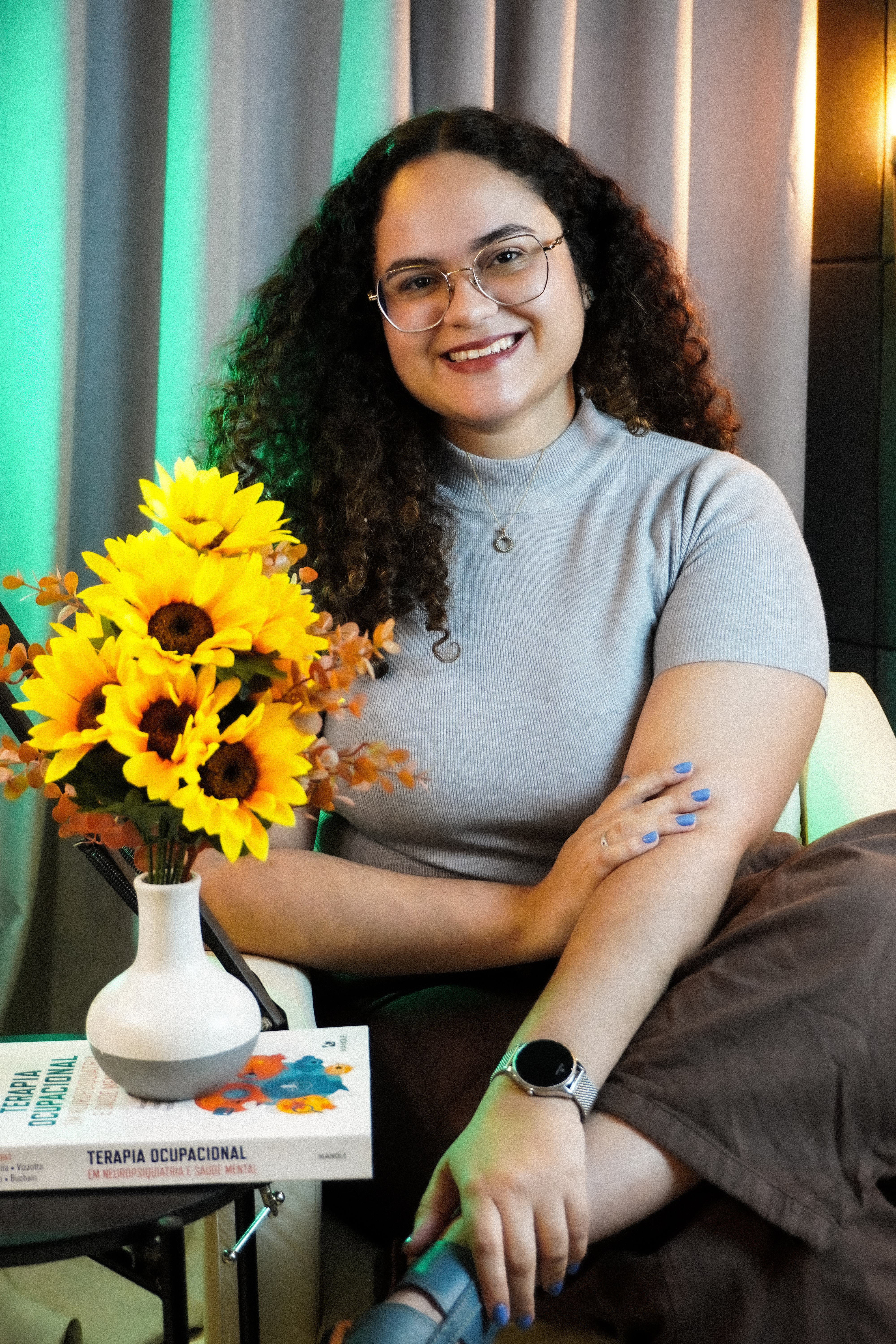 Brenda Ribeiro, terapeuta ocupacional, sentada com livro de Terapia Ocupacional e girassóis ao lado.